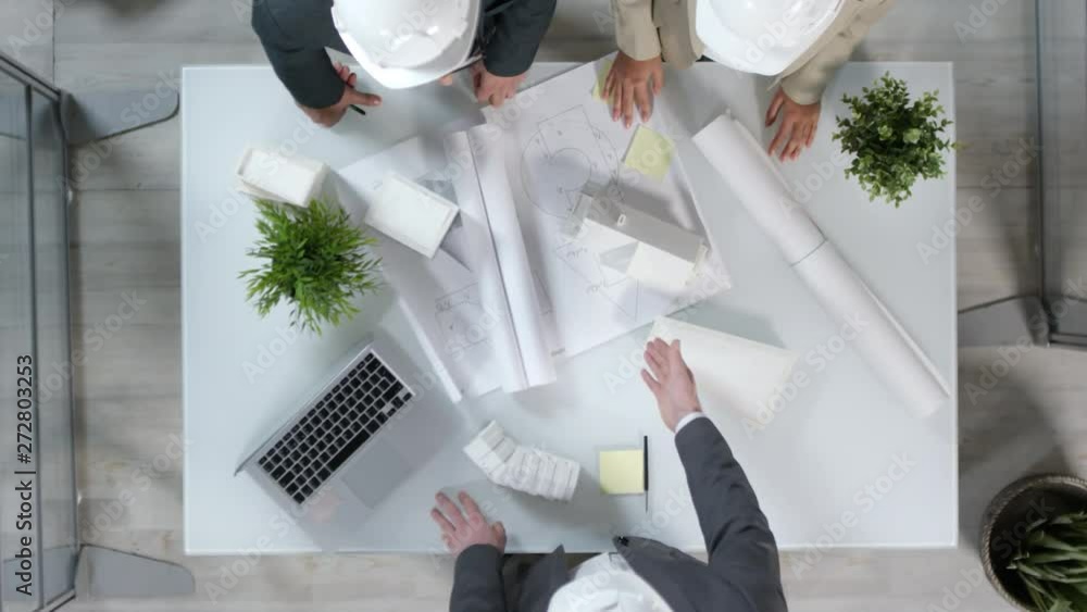 Directly above view of three male and female builders in white safety helmets standing at table, having conversation about new project and then shaking hands Directly above view of three male and female builders in white safety helmets standing at table, having conversation about new project and then shaking hands