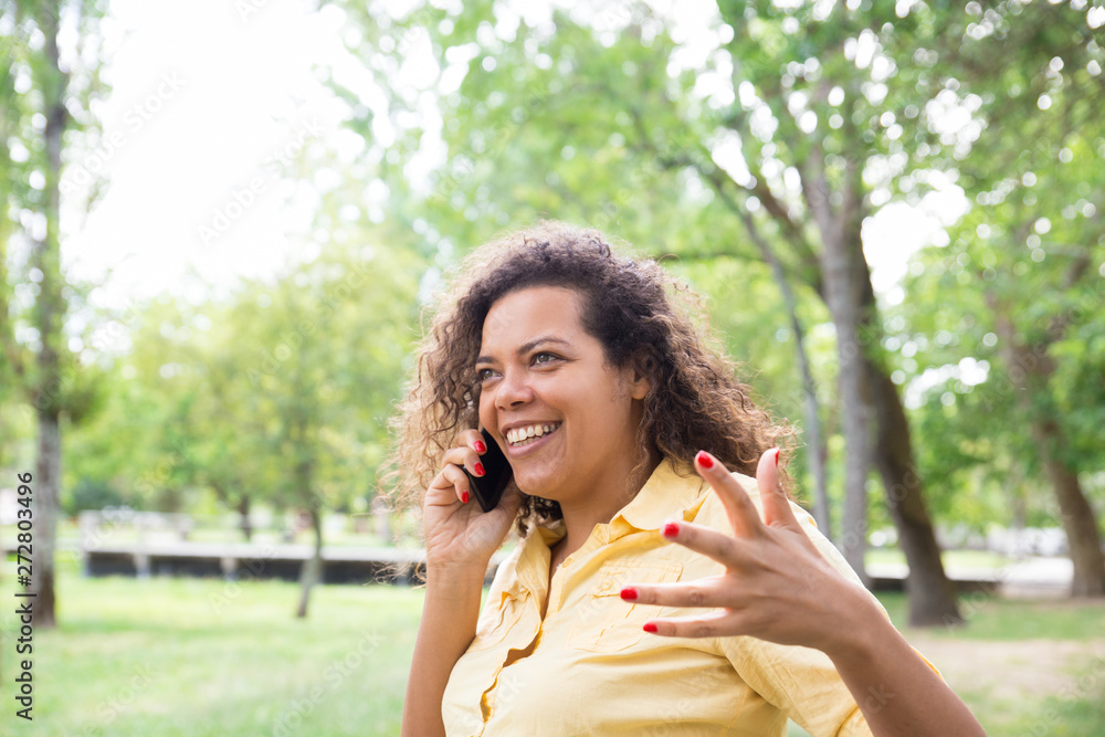 Smiling woman talking on phone and gesturing in city park. Young lady standing with blurred trees and lawn in background. Nature and communication concept. Smiling woman talking on phone and gesturing in city park. Young lady standing with blurred trees and lawn in background. Nature and communication concept.