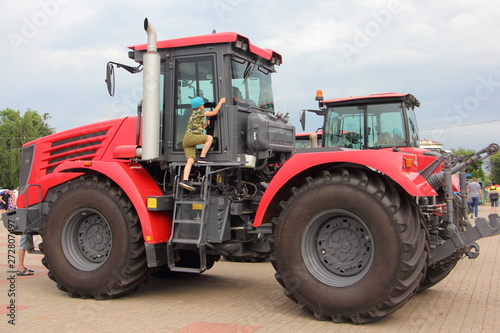 The boy climbs the stairs to the cockpit of a large new red tractor with big wheels at the exhibition of agricultural machinery