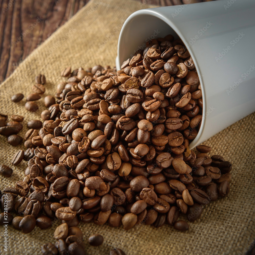 Fototapeta premium Coffee beans overflowing from a white coffee cup placed on a sackcloth and a black background.