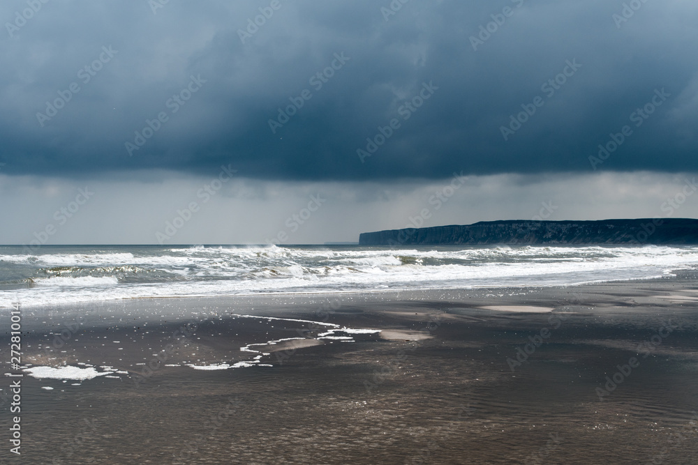 dark storm clouds above a flat sandy beach with white waves and sky reflecting in wet sand at Bempton Cliffs, UK in the background, depicting low colour palette, sun shining onto the sea from behind