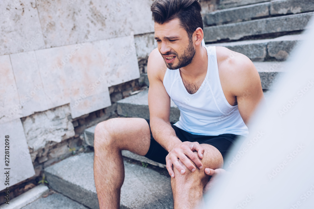 selective focus of handsome sportsman suffering from hurt while sitting on stairs and touching injured knee selective focus of handsome sportsman suffering from hurt while sitting on stairs and touching injured knee