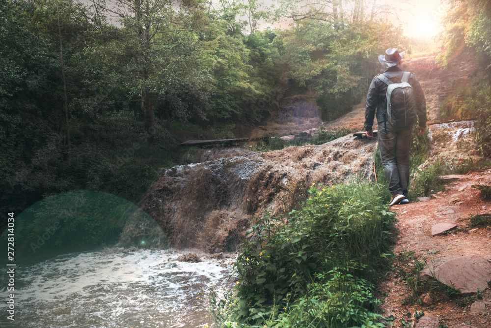 A man, a traveler in a leather jacket and a cowboy hat and backpack. Large full-flowing waterfall with dirty water, a journey, copy space. Travel, holidays, advanture. Dzhurinsky waterfall, Ukraine