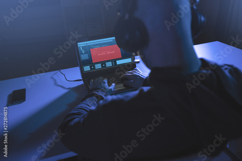 cybercrime, hacking and technology concept - male hacker in headphones with access denied message on laptop computer screen using virus program for cyber attack in dark room