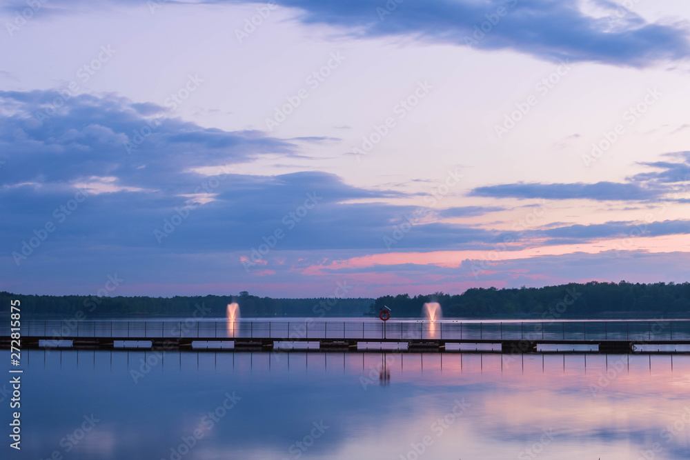 Colorful landscape with a wooden pier across the lake and two illuminated fountains under rhe blue cloudy sky in the evening. Reflection in water.