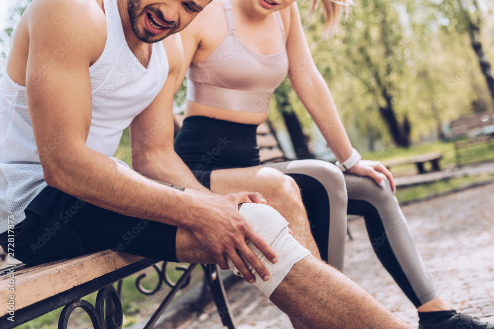 Obraz premium cropped shot of sportsman suffering from pain in injured knee while sitting on bench near young sportswoman