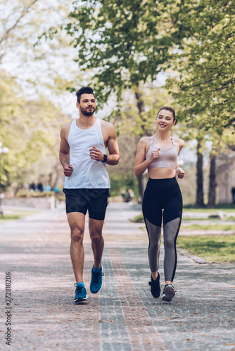 smiling sportsman and sportswoman running along wide alley in park