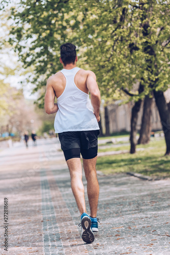 back view of young sportsman running along wide alley in park