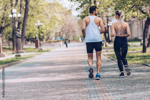 back view of man and woman in sportswear running along park alley