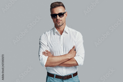 Perfect style. Handsome young man looking at camera and keeping arms crossed while standing against grey background