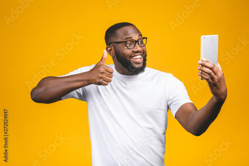 Portrait of cheerful afro american guy on holiday, isolated on yellow background, in casual wear, using veb cam on his tablet, enjoying, smiling. Thumbs up.