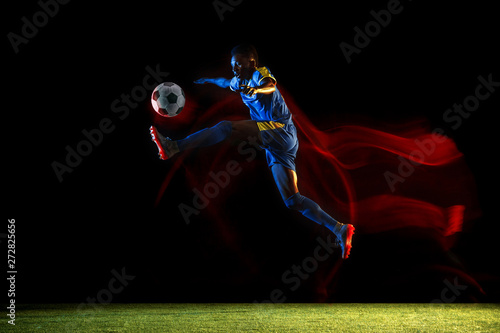 Young african-american male football or soccer player in sportwear and boots kicking ball for the goal in mixed light on dark background. Concept of healthy lifestyle, professional sport, hobby. © master1305