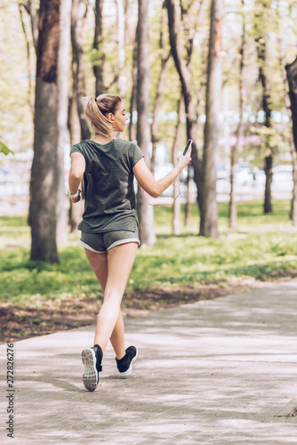 back view of young woman holding smartphone and listening music in earphones while jogging in park