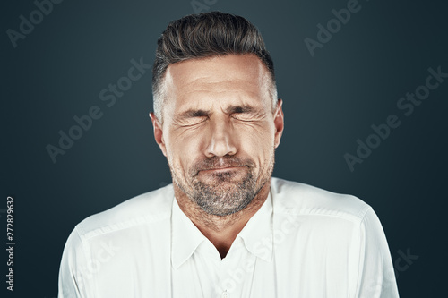 Grimacing. Handsome young man keeping eyes closed and inflating cheeks while standing against grey background photo
