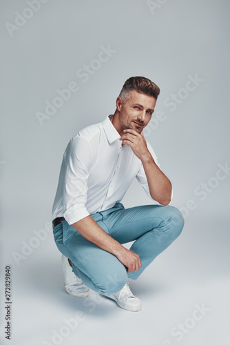 Male beauty. Handsome young man looking at camera and smiling while crouching against grey background photo