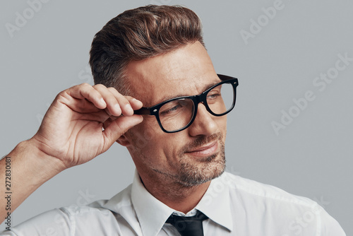 In his style. Good looking young man adjusting eyewear and smiling while standing against grey background