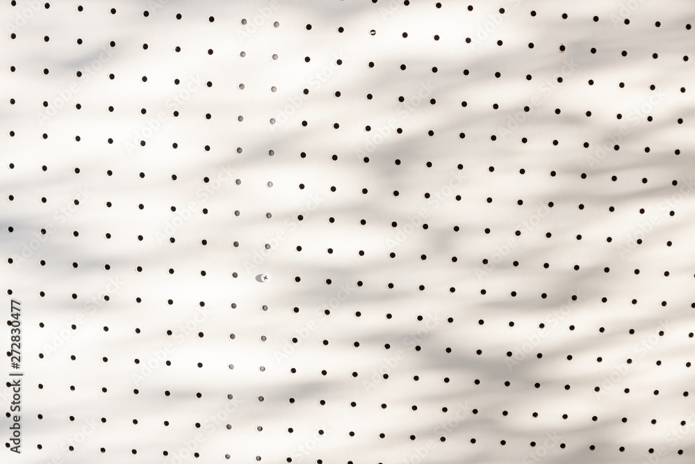 orderly dot or holes rows and columns on white pegboard wall with shadow of leaves and branch tree.