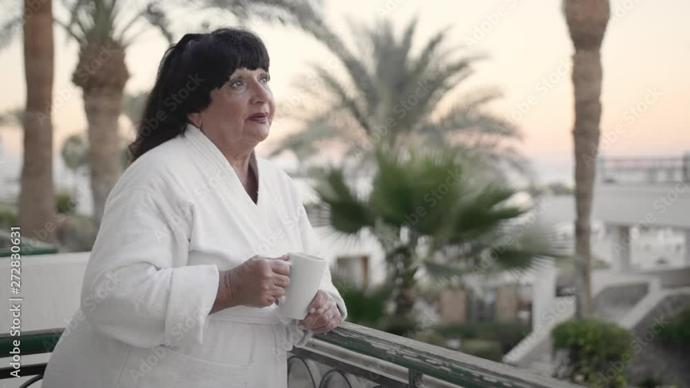 Caucasian senior woman in the morning drinking coffee on the hotel terrace in a white terry robe at a tropical resort. Against the backdrop of palm trees and the sea. Slow Motion, closeup. Caucasian senior woman in the morning drinking coffee on the hotel terrace in a white terry robe at a tropical resort. Against the backdrop of palm trees and the sea. Slow Motion, closeup.