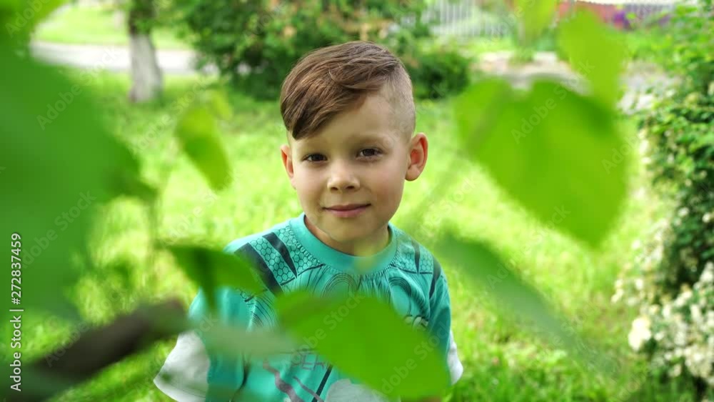 Serious boy in bright green garden. Adorable cute kid with stylish hair dress standing in bright greenery in street Serious boy in bright green garden. Adorable cute kid with stylish hair dress standing in bright greenery in street