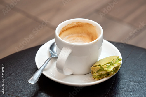 Half full cup of espresso macchiato with metal spoon and half eaten pistachio macaron on saucer. Black leather table.