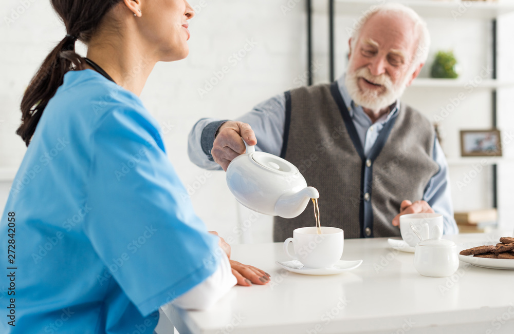 Happy and smiling senior man, pouring tea in white cup, sitting on kitchen at home Happy and smiling senior man, pouring tea in white cup, sitting on kitchen at home
