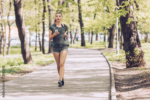 attractive young woman holding smartphone and listening music in earphones while jogging in park