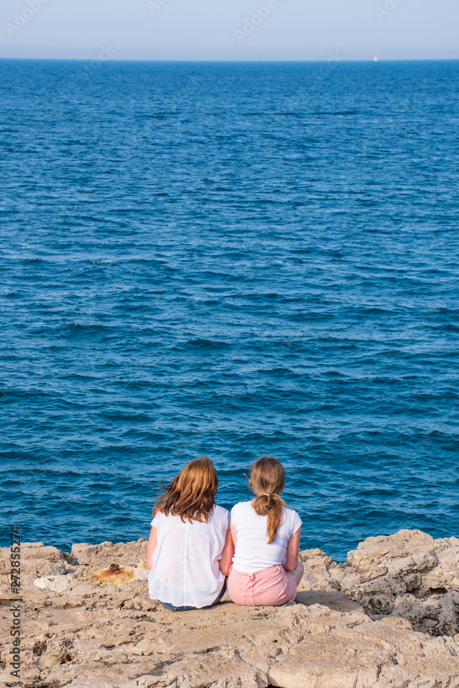 Fototapeta premium A couple of red hair girls, mother and daughter, sisters or friends standing on the rocks and looking at the blue sea, vertical