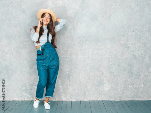 Young beautiful woman speaking on phone. Trendy girl in casual summer overalls clothes and hat. Funny and positive female posing near gray wall in studio