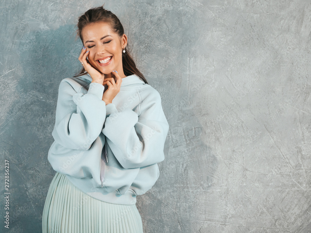 Young beautiful smiling woman looking at camera. Trendy girl in casual summer hoodie and skirt clothes. Funny and positive female posing near gray wall in studio Young beautiful smiling woman looking at camera. Trendy girl in casual summer hoodie and skirt clothes. Funny and positive female posing near gray wall in studio