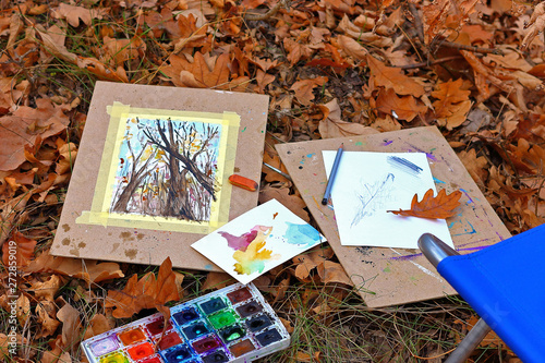 Watercolor paints, pencil and watercolor drawing on a pad lie on autumn brown fallen leaves