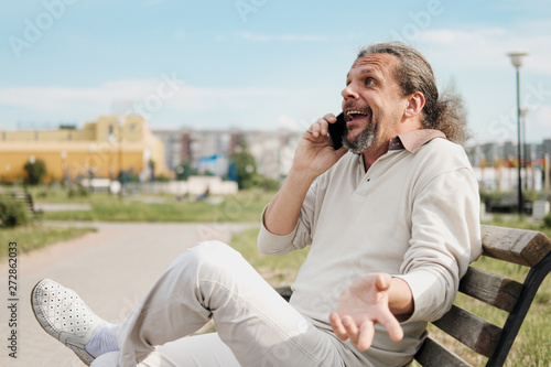 An elderly handsome man with long hair in the tail emotionally speaks on a cell phone in a park on a bench. © spaskov
