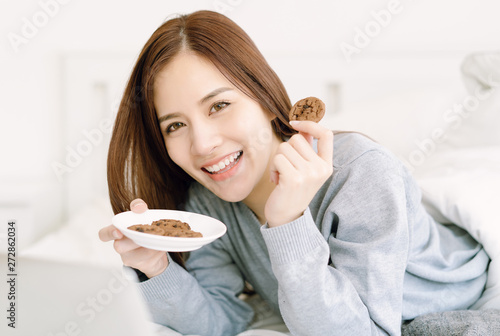 Portrait of beautiful young Asian woman wearing sweater eating cookie in bed at home bedroom, looking at camera. Relax and lifestyle Concept.