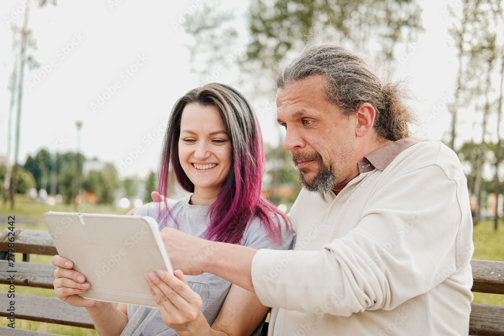 An elderly handsome man with long hair in the tail and a young beautiful woman are sitting on a park bench and using a tablet. Father and daughter relationship. An elderly handsome man with long hair in the tail and a young beautiful woman are sitting on a park bench and using a tablet. Father and daughter relationship.