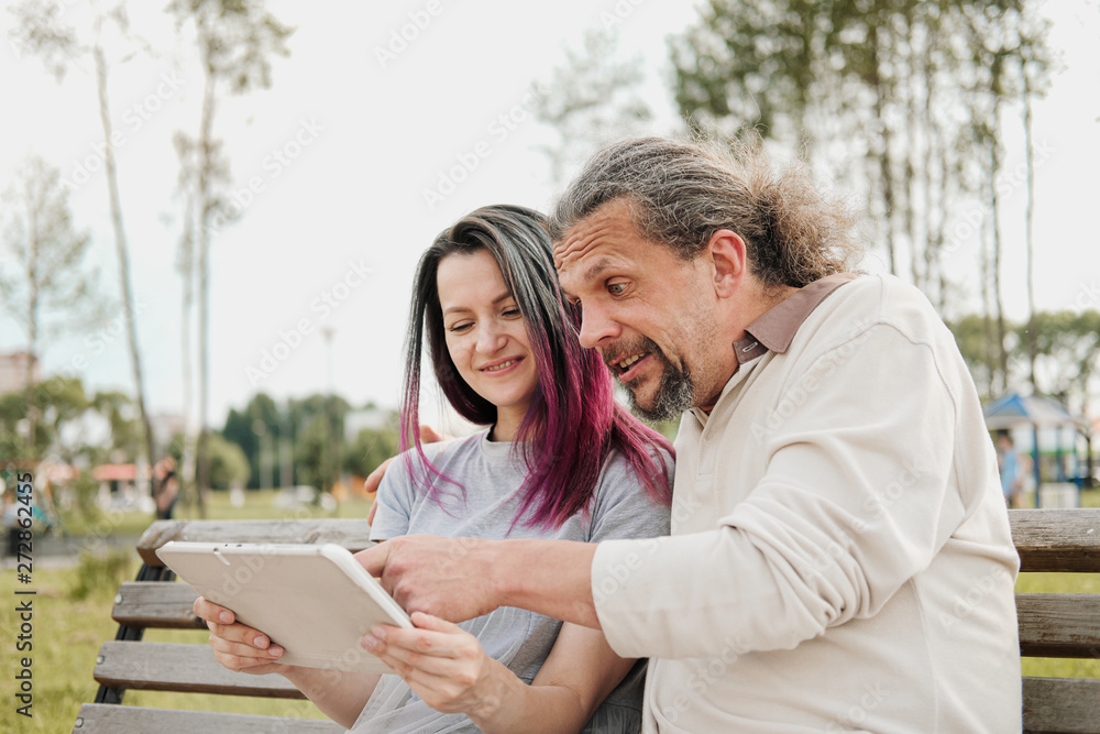 An elderly handsome man with long hair in the tail and a young beautiful woman are sitting on a park bench and using a tablet. Father and daughter relationship. An elderly handsome man with long hair in the tail and a young beautiful woman are sitting on a park bench and using a tablet. Father and daughter relationship.