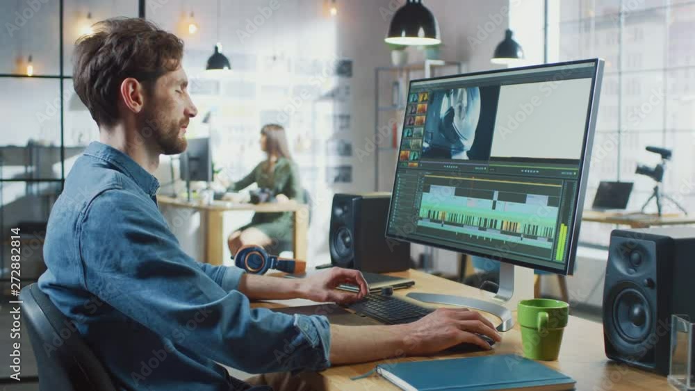 Male Video Editor with Beard and Jeans Shirt Works with Footage on His Personal Computer with Big Display. He Works in a Cool Office Loft. Other Female Creative Colleague Walks in the Background. Male Video Editor with Beard and Jeans Shirt Works with Footage on His Personal Computer with Big Display. He Works in a Cool Office Loft. Other Female Creative Colleague Walks in the Background.