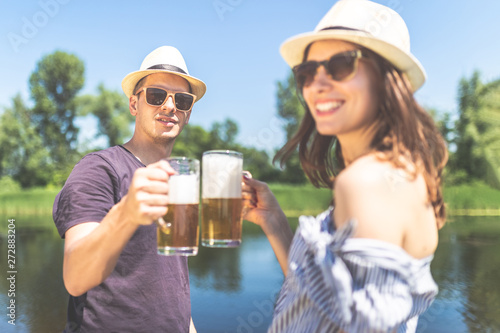Young cheerful hipster couple with beer mugs against beautiful lake in the background.