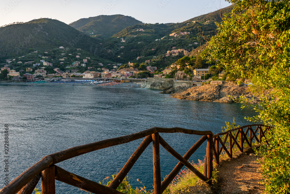 Mountain path secured by a wooden fence, leading to the Mediterranean sea. In the distance, the beach and green hills with colorful houses of Bonassola, Liguria, Italy. Mountain path secured by a wooden fence, leading to the Mediterranean sea. In the distance, the beach and green hills with colorful houses of Bonassola, Liguria, Italy.