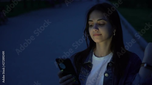 postive young girl using smartphone on the low-light street in the park in the evening