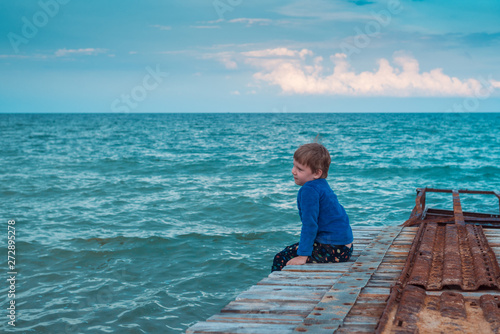 A little boy is sitting on the pier at sea.