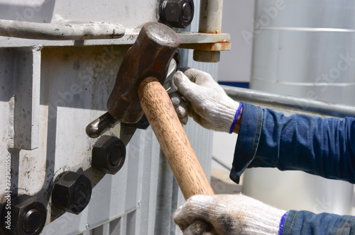 Workers use a wrench to loosen the nut by using a large hammer.  To open the metal manhole. The manhole is position overhead.