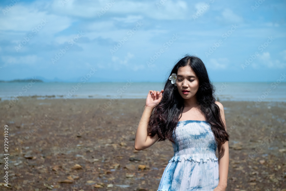 Asian girl wear Indigo dyed cotton long skirt,smiling and flower tucked behind her ear on the beach,blue sky and sea,orange light flare. Asian girl wear Indigo dyed cotton long skirt,smiling and flower tucked behind her ear on the beach,blue sky and sea,orange light flare.