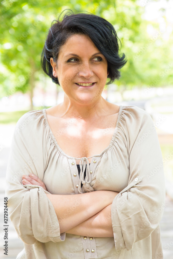 Happy black-haired woman standing for camera with arms crossed. Middle aged Caucasian lady posing in park, looking away and smiling. Outdoor female portrait concept Happy black-haired woman standing for camera with arms crossed. Middle aged Caucasian lady posing in park, looking away and smiling. Outdoor female portrait concept