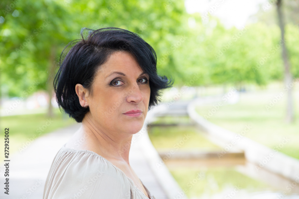 Serious confident woman in casual posing while walking in park. Closeup of middle aged Caucasian lady turning face to camera. Outdoor female portrait concept Serious confident woman in casual posing while walking in park. Closeup of middle aged Caucasian lady turning face to camera. Outdoor female portrait concept