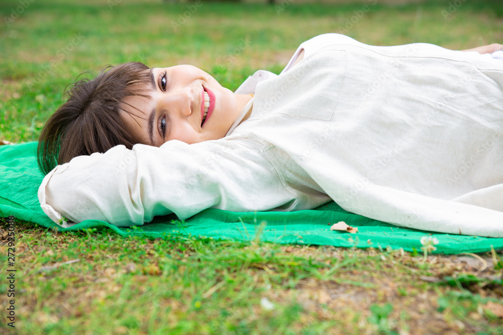 Happy pretty young woman lying and relaxing on lawn. Lady looking at camera and resting on blanket on ground. Rest and nature concept. Side view. Happy pretty young woman lying and relaxing on lawn. Lady looking at camera and resting on blanket on ground. Rest and nature concept. Side view.