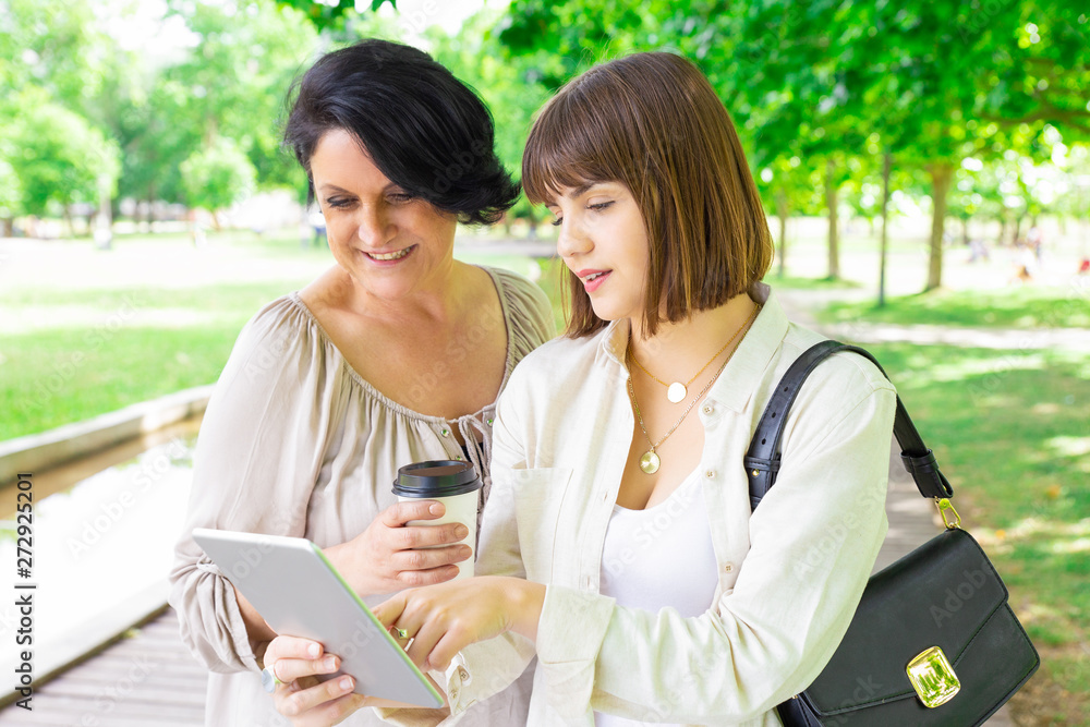Positive young woman and her mother using tablet in park. Middle-aged lady and her daughter standing and relaxing with trees in background. Family and communication concept. Front view. Positive young woman and her mother using tablet in park. Middle-aged lady and her daughter standing and relaxing with trees in background. Family and communication concept. Front view.