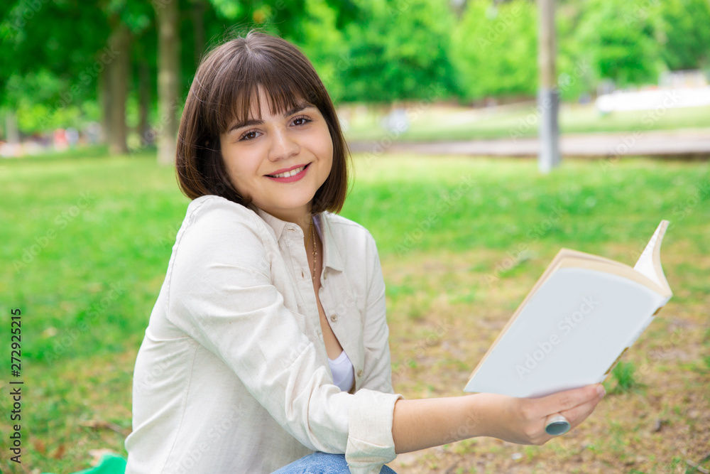 Positive young woman sitting and reading book on lawn. Pretty lady looking at camera and sitting on ground with trees in background. Education and nature concept.