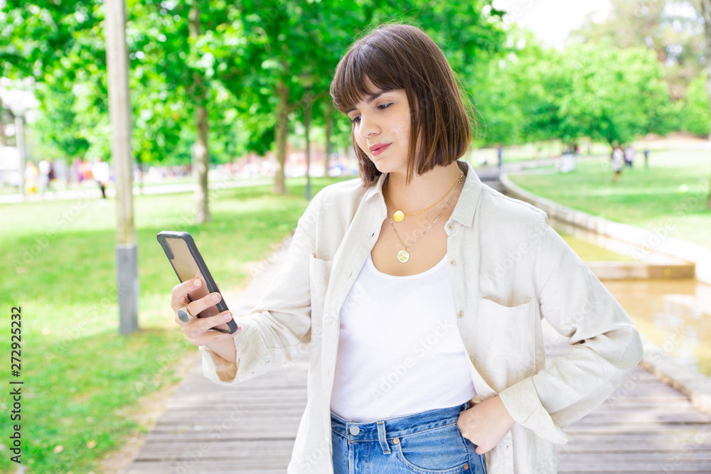 Serious pretty woman using smartphone in city park. Young lady standing with trees and walkway in background. Communication and summer concept. Front view. Serious pretty woman using smartphone in city park. Young lady standing with trees and walkway in background. Communication and summer concept. Front view.