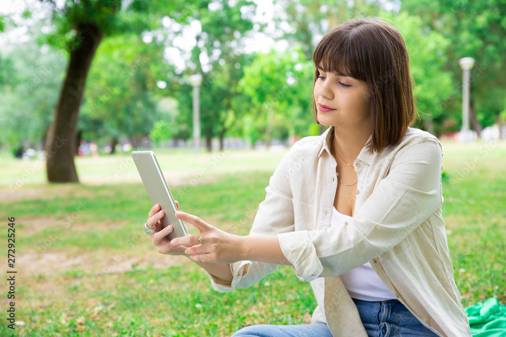 Serious pretty woman using tablet and sitting on lawn. Young lady sitting on ground with trees in background. Technology and relaxation concept.