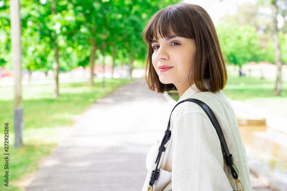 Serious pretty young woman walking in city park. Lady looking away with trees and pavement in background. Leisure and nature concept. Side view. Serious pretty young woman walking in city park. Lady looking away with trees and pavement in background. Leisure and nature concept. Side view.