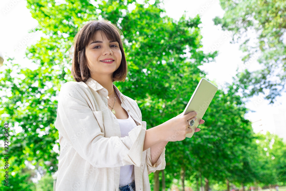 Smiling young woman reading news on tablet and standing in park. Pretty lady wearing casual shirt and looking at camera with green trees in background. Technology and summer concept. Smiling young woman reading news on tablet and standing in park. Pretty lady wearing casual shirt and looking at camera with green trees in background. Technology and summer concept.